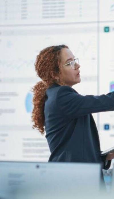 Hispanic Female Senior Data Scientist Reviewing Reports Of Risk Management Department On Big Digital Screen In Monitoring Room. Diverse Consulting Company Employees Working Behind Desktop Computers.